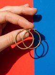 Hand houdt een paar goudkleurige, ronde hoepeloorbellen vast tegen een achtergrond van rood en blauw.