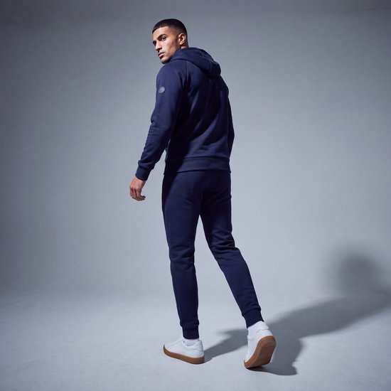 Model draagt een marineblauw trainingspak met Mario Russo-embleem op de mouw, gecombineerd met witte sneakers.