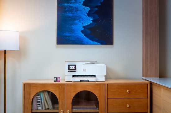Witte HP Envy All-in-One printer met kleurenscherm op een houten dressoir, naast twee inktcartridges.