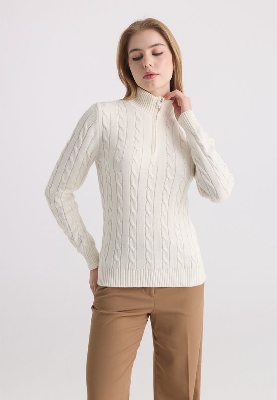 Femme aux longs cheveux blonds, portant un pull écru à col roulé torsadé et zip, avec un pantalon marron. Vue de côté.