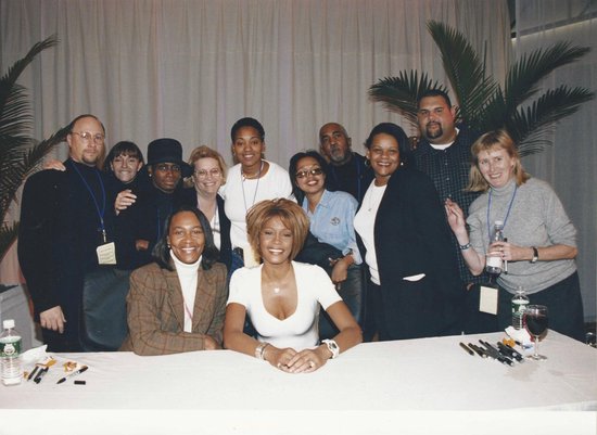 Whitney Houston met een groep mensen achter een tafel, ze zit vooraan.
