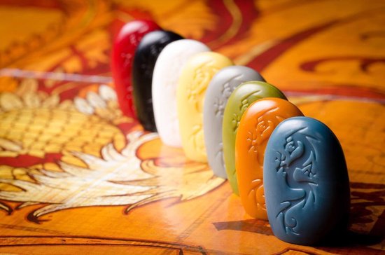 Zeven gekleurde Tsuro-stenen (rood, zwart, wit, geel, grijs, groen, oranje, blauw) op een speltafel.