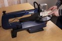 Blauwe FERM figuurzaagmachine op een houten tafel, met een stuk hout in bewerking. Iemand heeft handen op het hout.