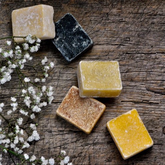 Vijf vierkante amberblokjes in geuren van amber, vanille, witte jasmijn, zwarte musk en witte musk, met witte bloesems op een houten ondergrond.
