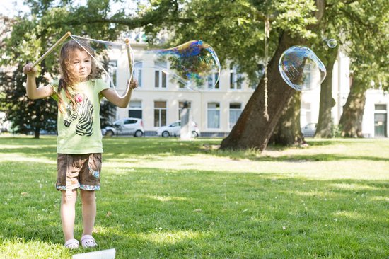 Meisje in geel shirt en bruine shorts blaast een grote zeepbel in de tuin.