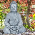 Lichtgrijs zittend Boeddhabeeld in een tuin met Japanse esdoorn en stenen.