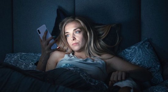 Een blonde vrouw met blauwe ogen ligt 's nachts in bed met haar smartphone.