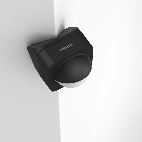 Zwarte Philips Hue buitenbewegingssensor met 'PHILIPS' logo, in een hoek op een witte muur.