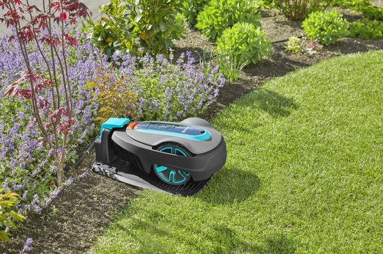 Grijze robotmaaier met blauwe en oranje accenten, geparkeerd op een laadstation aan de rand van een bloembed.