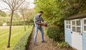 Man met heggenschaar naast een blauw tuinhuisje.