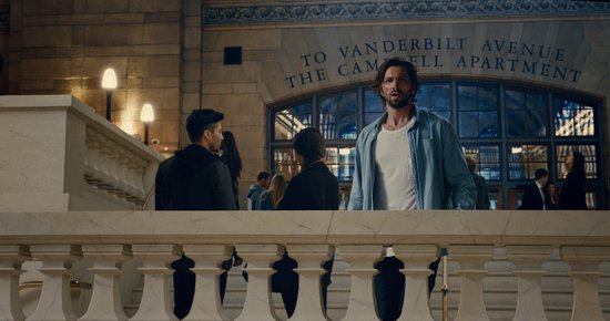 Een man met een lichtblauwe jas en wit T-shirt staat in een hal met een tekst aan de muur: 'To Vanderbilt Avenue The Campbell Apartment'.
