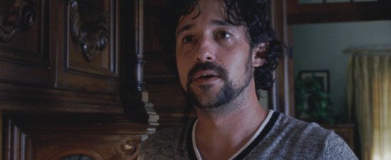Man met donker haar en baard draagt een grijs shirt met witte bies en kijkt met open mond naar voren.