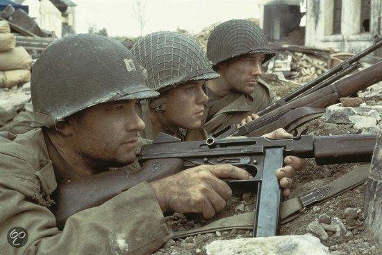 Drie soldaten met helmen en wapens richten vanuit een verwoeste omgeving. Tom Hanks is vooraan.