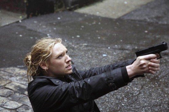 Een vrouw met blond haar in een zwarte jas houdt een pistool vast in de regen.