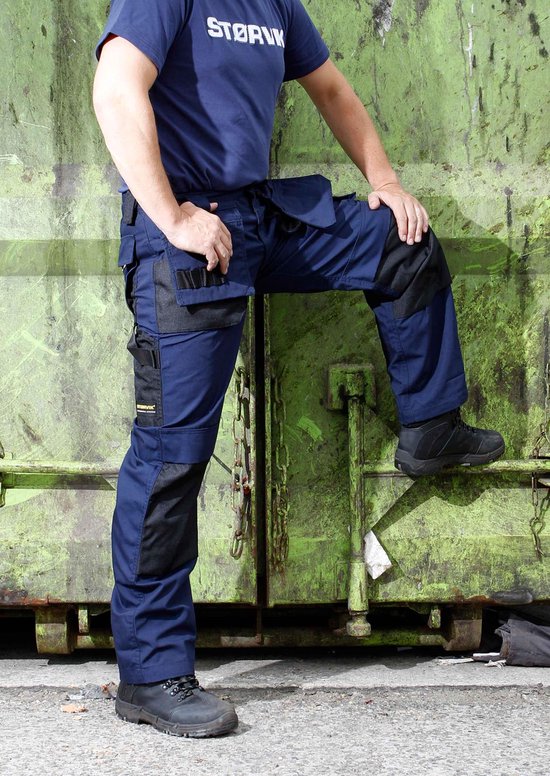 Marineblauwe werkbroek met zwarte accenten op de zakken en knieën, gedragen door een model met de voet op een container.