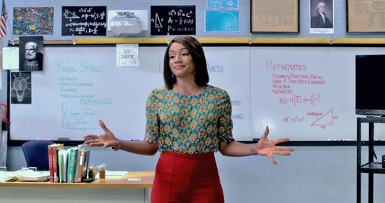 Een vrouw in een kleurrijk shirt met patronen en rode broek staat in een klaslokaal voor een whiteboard.