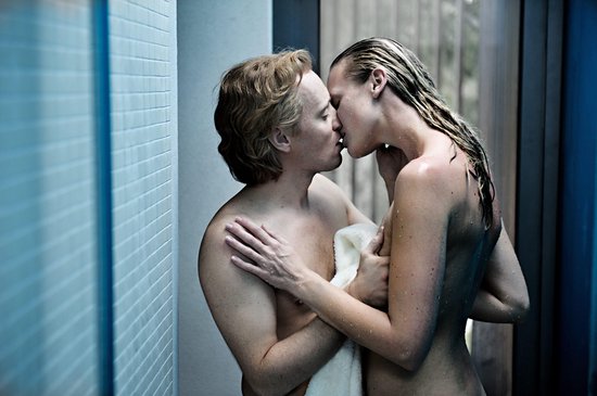 Man en vrouw zoenen in de douche, vrouw omhelsd door man met handdoek.