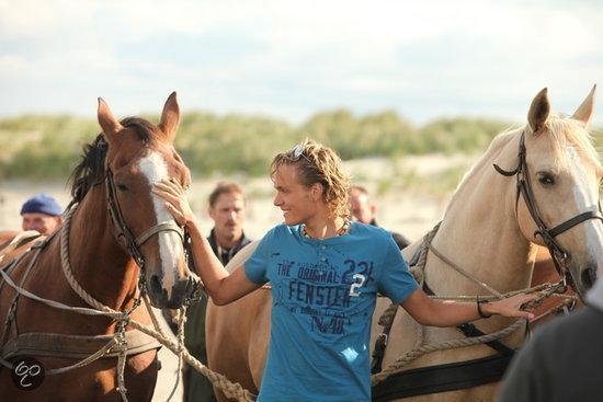 Jonge man met blond haar en zonnebril raakt twee paarden aan op het strand.