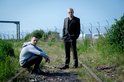Twee mannen op spoorweg: één in trainingspak, één in pak, in een landelijke omgeving.