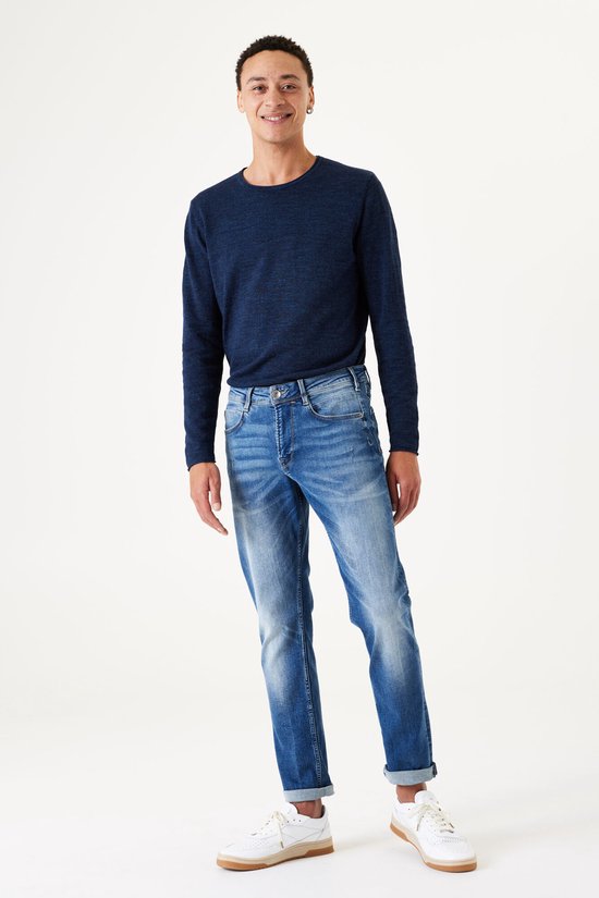 Man in een donkerblauwe trui en een lichtblauwe slim fit jeans, staand.