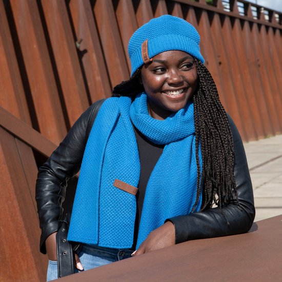 Vrouw met gevlochten haar, blauwe beanie en sjaal, draagt blauwe denim jas.