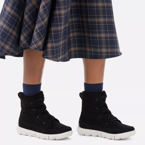 Zwarte Sorel snowboots met witte zolen en een teddy-achtige schacht. Gedragen met een geruite rok en blauwe sokken.