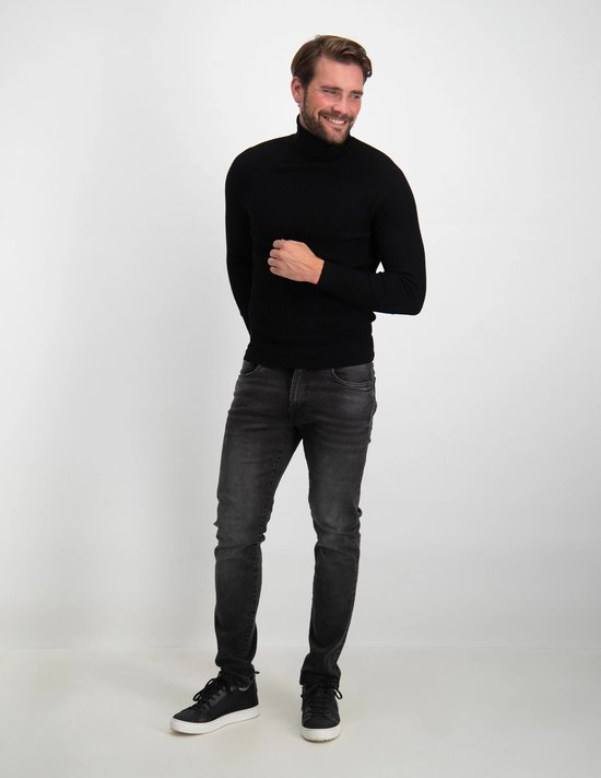 Man lach in zwarte coltrui en donkergrijze slim fit jeans, met zwarte sneakers.