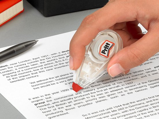 Main tenant un rouleau correcteur Pritt mini, l'appliquant sur du texte dans un document. Le rouleau est transparent avec des détails rouges.