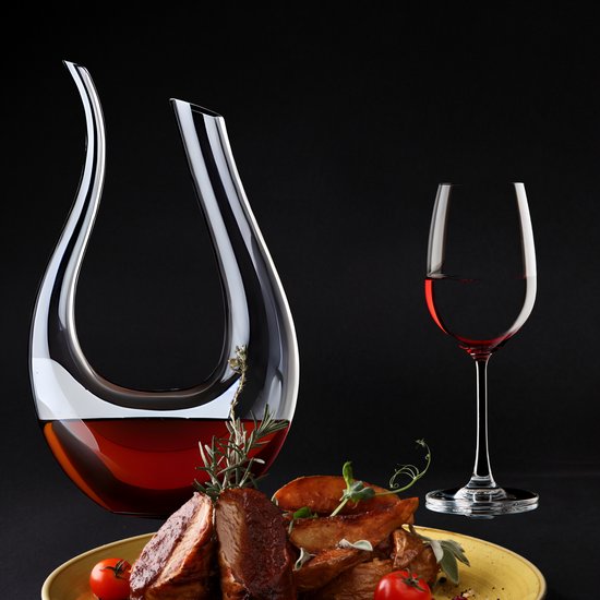 Une carafe à vin en verre avec un col courbé se trouve à côté d'un verre de vin rouge et d'une assiette de viande grillée et de pommes de terre.