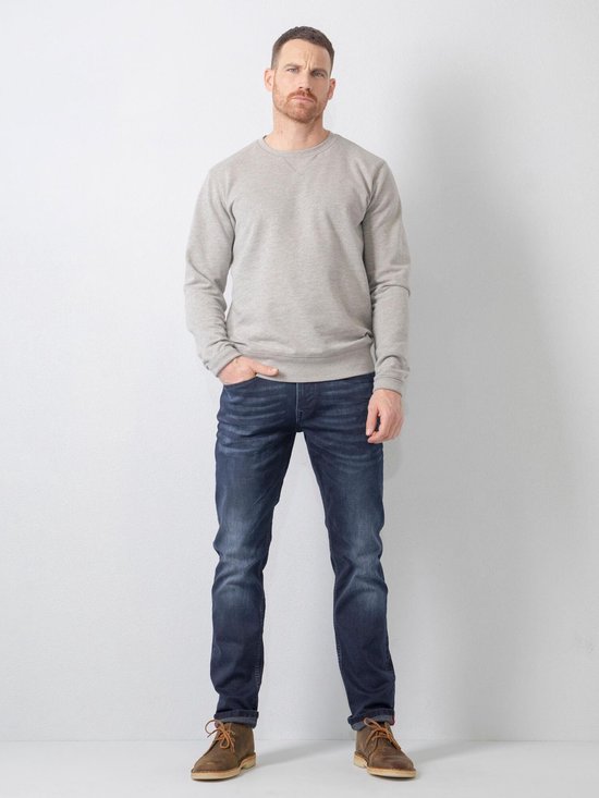 Man staat naar voren gericht, draagt een grijze trui en donkerblauwe jeans.