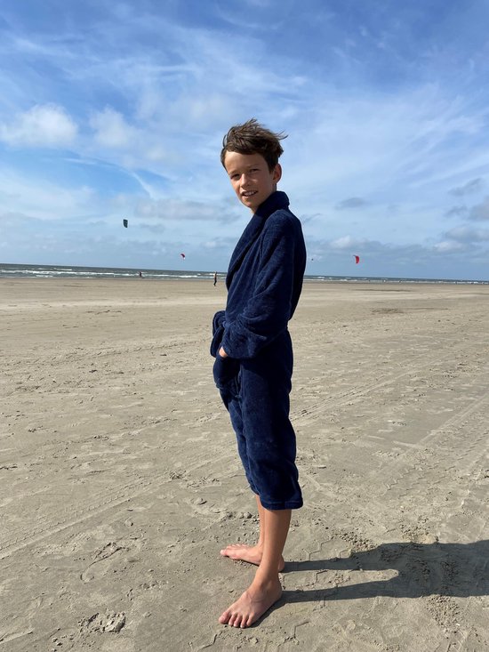 Jongen in een donkerblauwe fleeced badjas met ceintuur, staand op het strand.