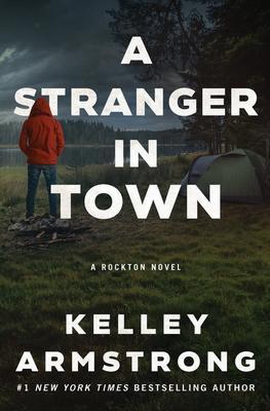 Boekomslag met de titel 'A Stranger in Town' van Kelley Armstrong. Een man in een rode hoodie staat met de rug naar de kijker, kijkend naar een meer en tent.