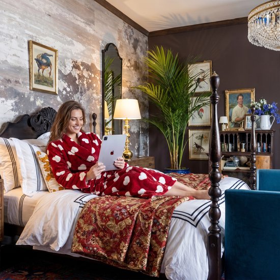 Een vrouw op een bed met een rode badjas met witte hartjes, kijkend naar een tablet.