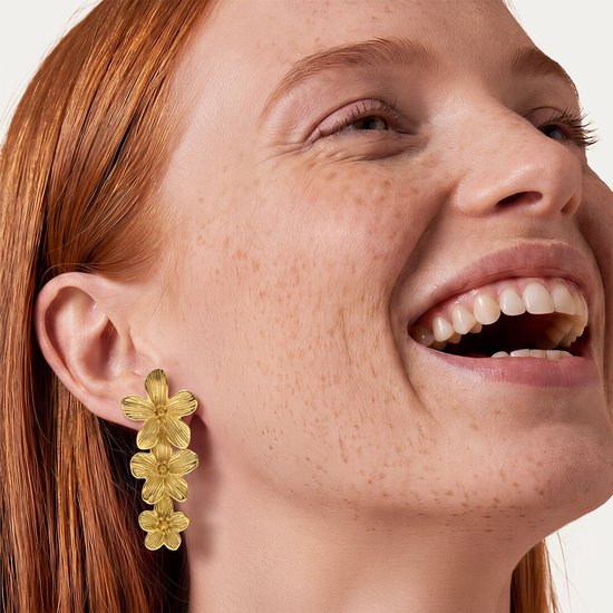 Gouden oorbellen met drie gestapelde bloemen, gedragen door een vrouw met rood haar en sproeten.