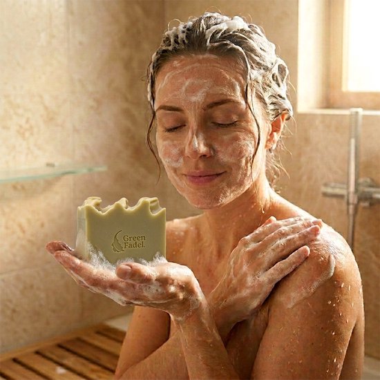 Vrouw met gesloten ogen in de douche, zeep in haar handen en gezicht. Ze houdt een groene "Green Fadel" zeep vast.
