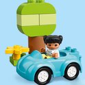 Blauwe LEGO Duplo auto met een meisje met zwarte staartjes erin, naast een boom met een gele bloem.