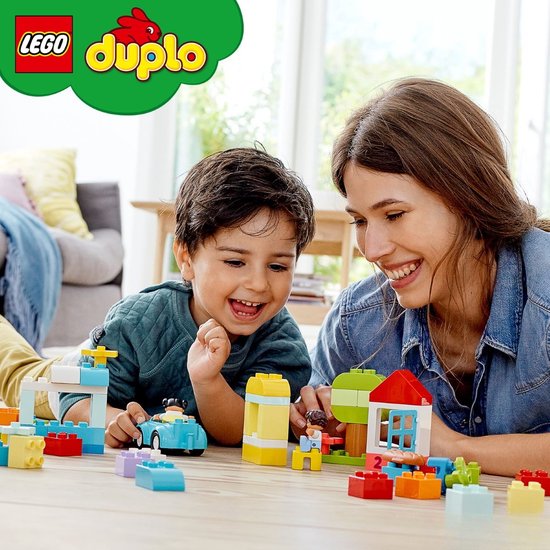 Moeder en kind spelen met kleurrijke LEGO Duplo stenen, waaronder een huis, auto en boom.