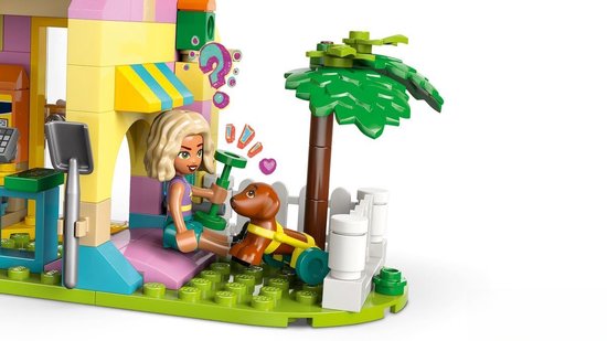 Gros plan d'une mini-poupée LEGO Friends tenant un chiot marron en laisse devant une animalerie rose et jaune.