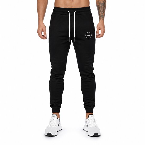 Man staand in zwarte trainingsbroek met witte veters, witte sneakers en MW logo.