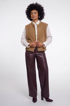 Model met krullend haar draagt bruin, gebreid vest met ronde hals, wit overhemd en bruine leren broek.