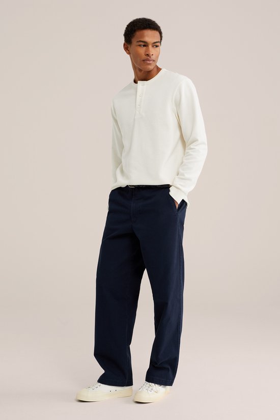 Model staat in een donkerblauwe chino en een wit longsleeve shirt.