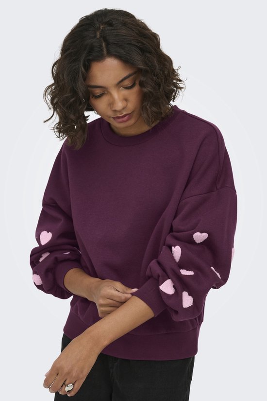 Donkerpaarse sweater met geborduurde lichtroze hartjes op de mouwen. Model met donkere krullen en huid, poseert.