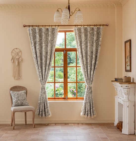Twee crèmekleurige gordijnen met bruine barokprint en franjes hangen voor een raam, naast een open haard en stoel.