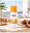 Eucerin zonnebrandcrème met SPF 50 in witte fles met oranje dop. De fles staat op een stenen tafel met een strooien hoed en zonnebril.