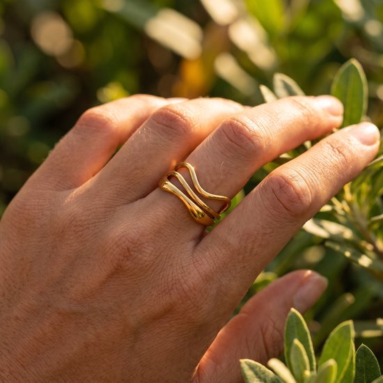 Gouden ring met golvende lijnen, gedragen op een hand tussen groen gebladerte.