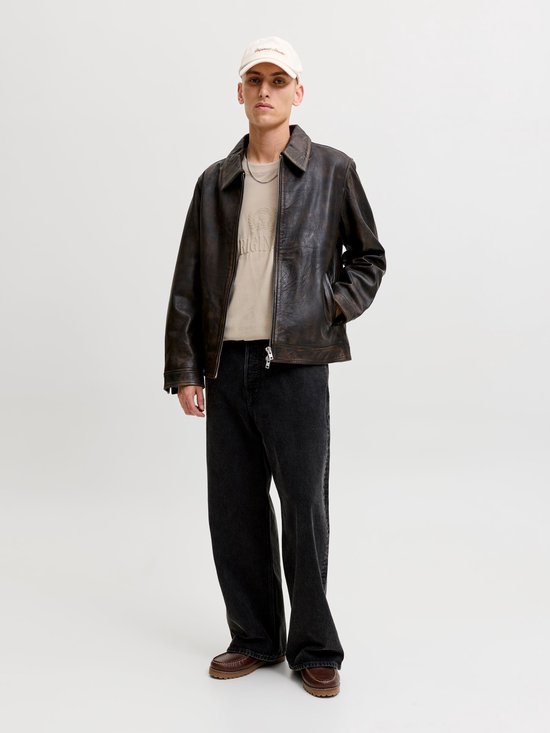 Man met beige T-shirt met "ORIGINALS" opdruk, leren jack, zwart jeans en pet.