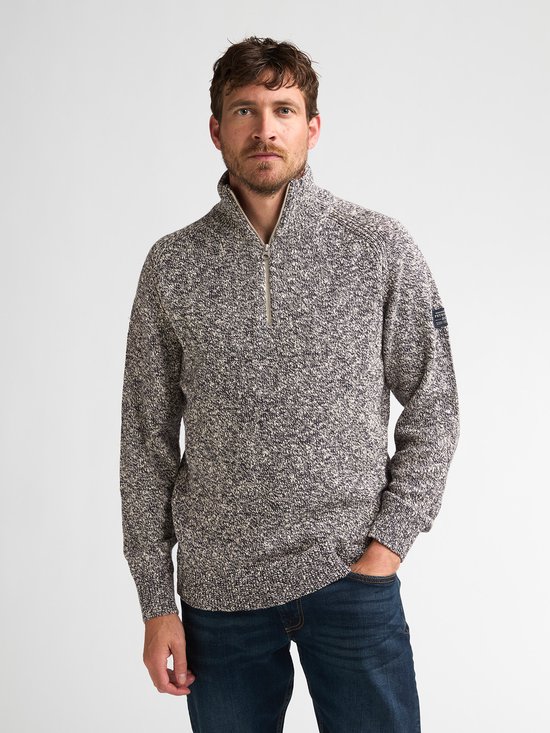 Man met gemêleerde blauw-witte trui met rits, logo op de mouw en donkere spijkerbroek.