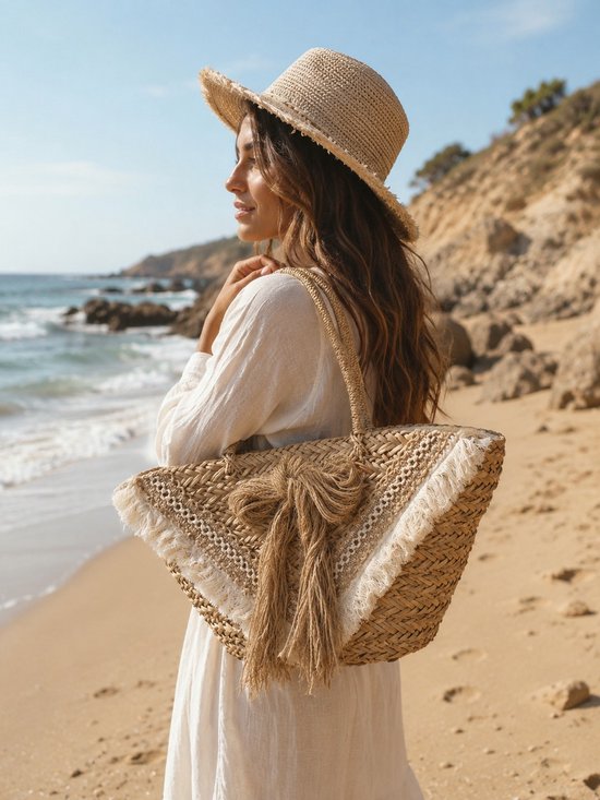 Une femme coiffée d'un chapeau de paille et vêtue d'une robe blanche porte un panier en paille à franges sur la plage.