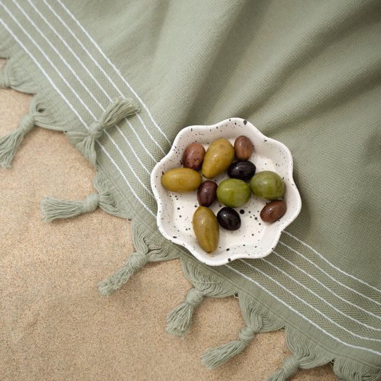 Een schaaltje olijven op een groene hamamdoek met witte strepen en franjes, op zand.
