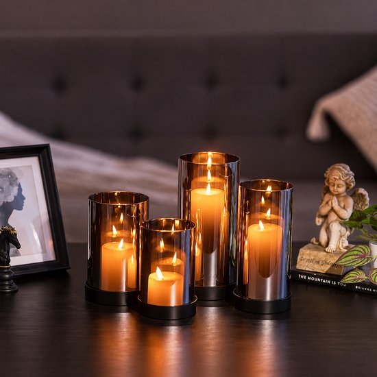 Vier brandende kaarsen in glazen windlichten op een donkere tafel, met een cherubijn en een ingelijste foto op de achtergrond.